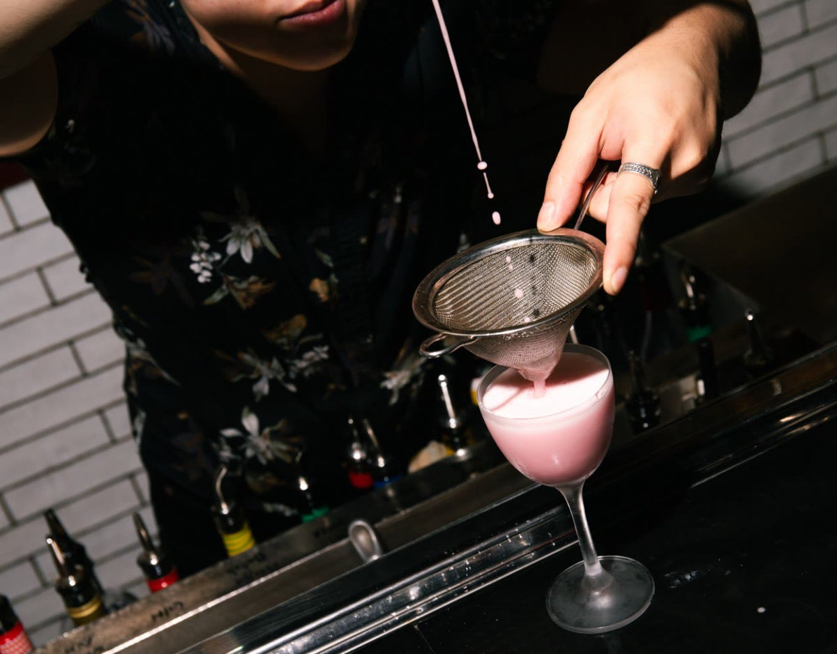 Cocktail Foamer serve — bartender double-straining a foam-topped pink cocktail into a coupe glass