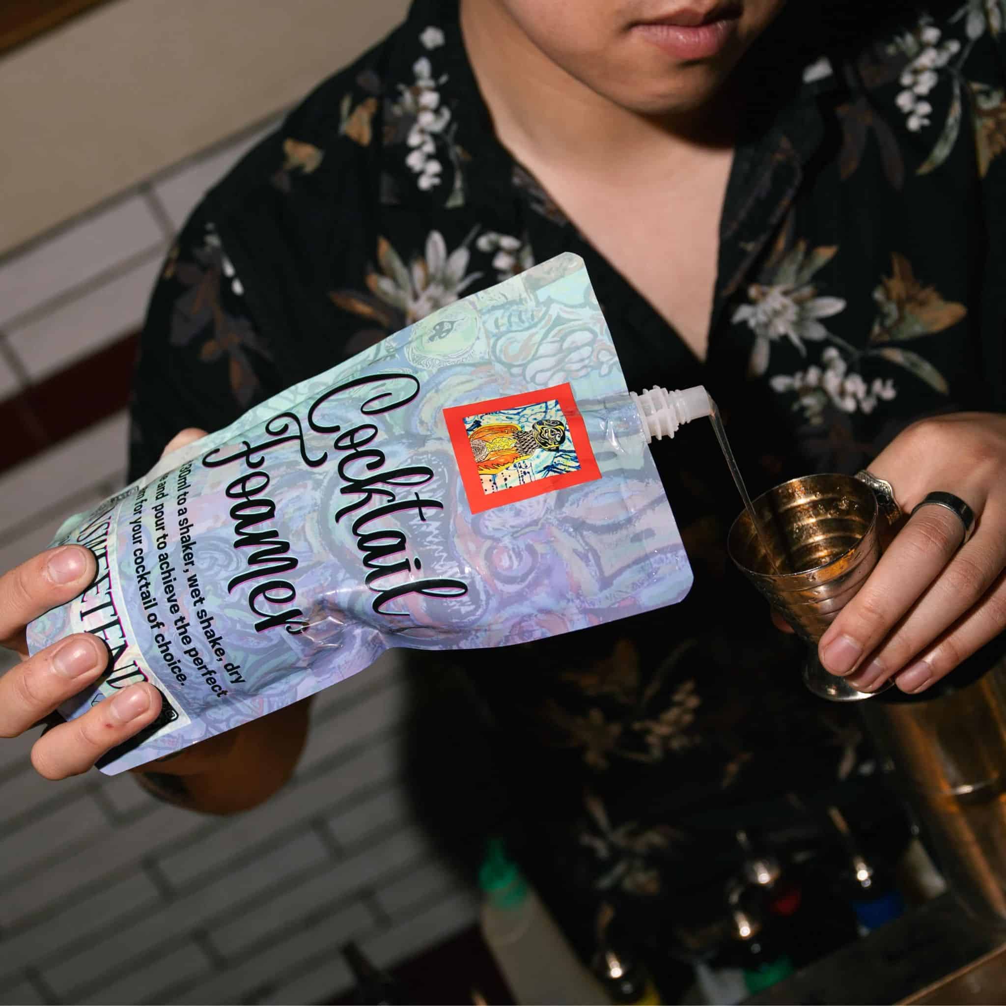 Person holding a Cocktail Foamer mixer packet.  pouring into a 75ml metal measuring jigger wearing a dark floral shirt. 