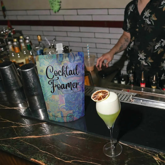 Cocktail Foamer 500mL matte-finish pouch displayed on a bar counter alongside a finished sour cocktail with thick white foam head, professional bar setting with warm ambient lighting