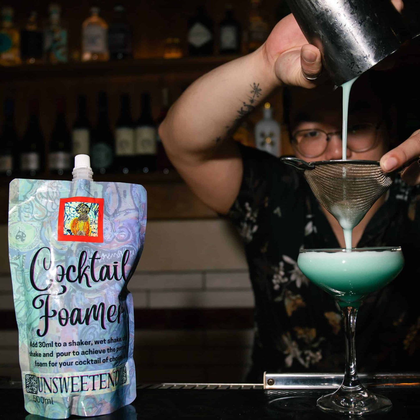 Person pouring / straining a blue cocktail from a pouch labeled 'Cocktail Foamer' into a glass.